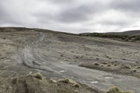 two motorcycles driving on dirt and sand near rocks and a hill and shrubs under cloudy skies