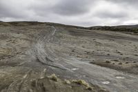 two motorcycles driving on dirt and sand near rocks and a hill and shrubs under cloudy skies