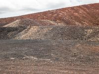 two motorcycles driving near a mountain of dirt and rocks near clouds in the background a red mountain top and a lone, mostly cloudy sky