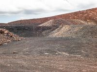 two motorcycles driving near a mountain of dirt and rocks near clouds in the background a red mountain top and a lone, mostly cloudy sky