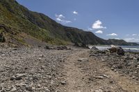 a rocky beach next to a cliff by the sea and a mountain range with green vegetation