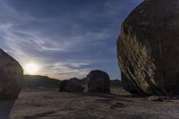 large rocks line a dirt field at sunset with the sun shining behind them, in a barren landscape