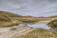 a dirt road surrounded by grass and dirt hills with a creek on one side of it