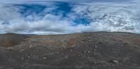 the view from a large grassy hill with rocks in the desert, with a very cloudy blue sky overhead