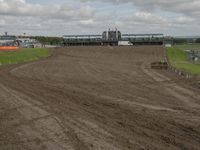 a dirt track with three horses in it and a large building in the background and clouds overhead