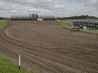 a dirt track with three horses in it and a large building in the background and clouds overhead