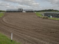 a dirt track with three horses in it and a large building in the background and clouds overhead