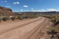 an empty dirt road in the desert near mountains and rocks with green bushes, grass, and dirt