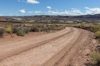 an empty dirt road in the desert near mountains and rocks with green bushes, grass, and dirt