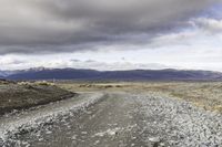 a gravel road next to grass and mountains under cloudy skies during the day in iceland