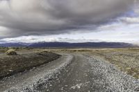 a gravel road next to grass and mountains under cloudy skies during the day in iceland