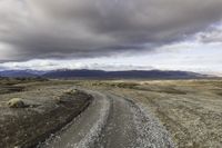 a gravel road next to grass and mountains under cloudy skies during the day in iceland