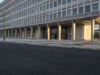 cars driving down a city street near a building with columns in front of it and people walking on the side of the road in front of it