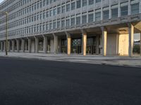 cars driving down a city street near a building with columns in front of it and people walking on the side of the road in front of it