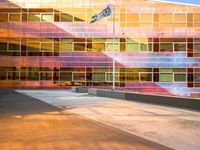 a man on a skateboard in front of a building with colorful glass walls and a flag pole