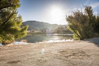 an empty dirt road with trees and a lake below them in front of the sun