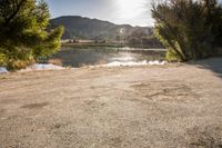 an empty dirt road with trees and a lake below them in front of the sun