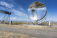 an old satellite mirror sitting on top of a dirt field near a blue sky and grass