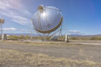 an old satellite mirror sitting on top of a dirt field near a blue sky and grass