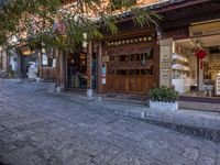 a stone street with plants and shops on either side of the road near a tree