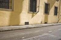 an old yellow building is on the side of the street and there are two windows, and a bicycle parked near it