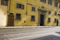 an old yellow building is on the side of the street and there are two windows, and a bicycle parked near it