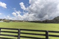 a field with a black wooden fence surrounded by clouds, and the sky is filled with clouds