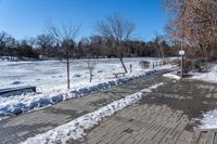 a walkway surrounded by snow next to a tree and bench in a park with clear skies