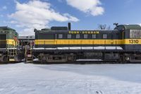 a yellow and black train with it's headlights on is in the snow with another locomotive behind it