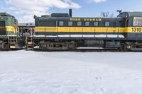 a yellow and black train with it's headlights on is in the snow with another locomotive behind it