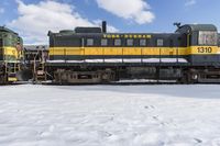 a yellow and black train with it's headlights on is in the snow with another locomotive behind it