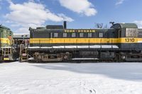 a yellow and black train with it's headlights on is in the snow with another locomotive behind it