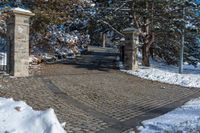 a driveway that has been decorated with stone blocks, pillars and trees with snow on the ground