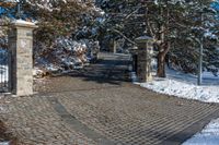 a driveway that has been decorated with stone blocks, pillars and trees with snow on the ground