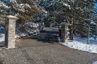 a driveway that has been decorated with stone blocks, pillars and trees with snow on the ground