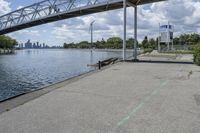 a metal bridge spanning over the water in a park area by a river and city skyline