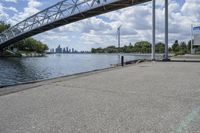 a metal bridge spanning over the water in a park area by a river and city skyline