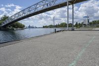 a metal bridge spanning over the water in a park area by a river and city skyline