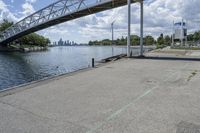 a metal bridge spanning over the water in a park area by a river and city skyline