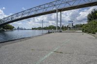 a metal bridge spanning over the water in a park area by a river and city skyline