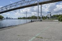 a metal bridge spanning over the water in a park area by a river and city skyline