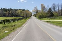 an empty country road surrounded by grass and fenced in field and trees in background