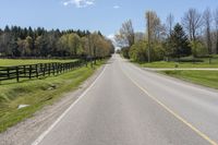 an empty country road surrounded by grass and fenced in field and trees in background