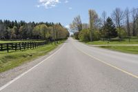 an empty country road surrounded by grass and fenced in field and trees in background