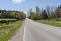 an empty country road surrounded by grass and fenced in field and trees in background