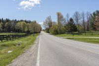 an empty country road surrounded by grass and fenced in field and trees in background