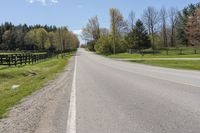 an empty country road surrounded by grass and fenced in field and trees in background