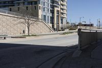 a city street lined with tall building next to an empty sidewalk and stone fence with benches near the sidewalk