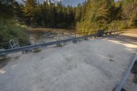 a road with the railing in front of it and water flowing in the background of trees