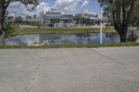 a blue sky with clouds and buildings in the background a pond is shown with the grass on one side and trees and some water and shrubs